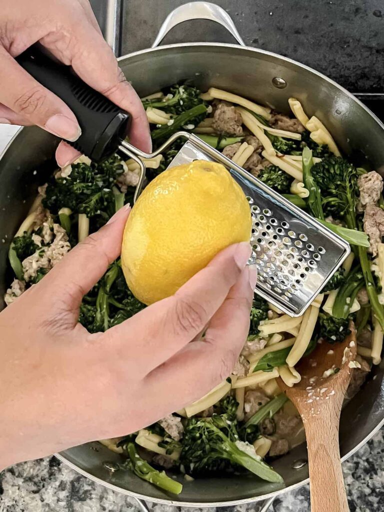 Brown hands zesting a lemon into a pot of casarecce with broccolini and Italian sausage.