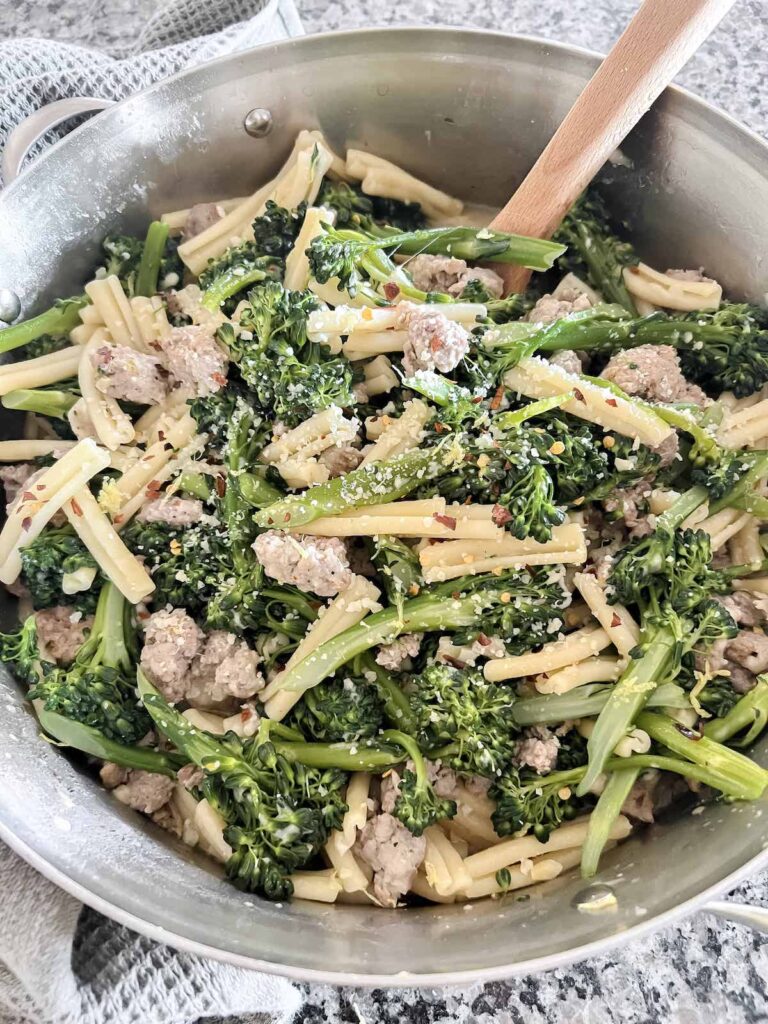 Casarecce with broccolini and Italian sausage in a stainless steel pot with a wooden spoon.