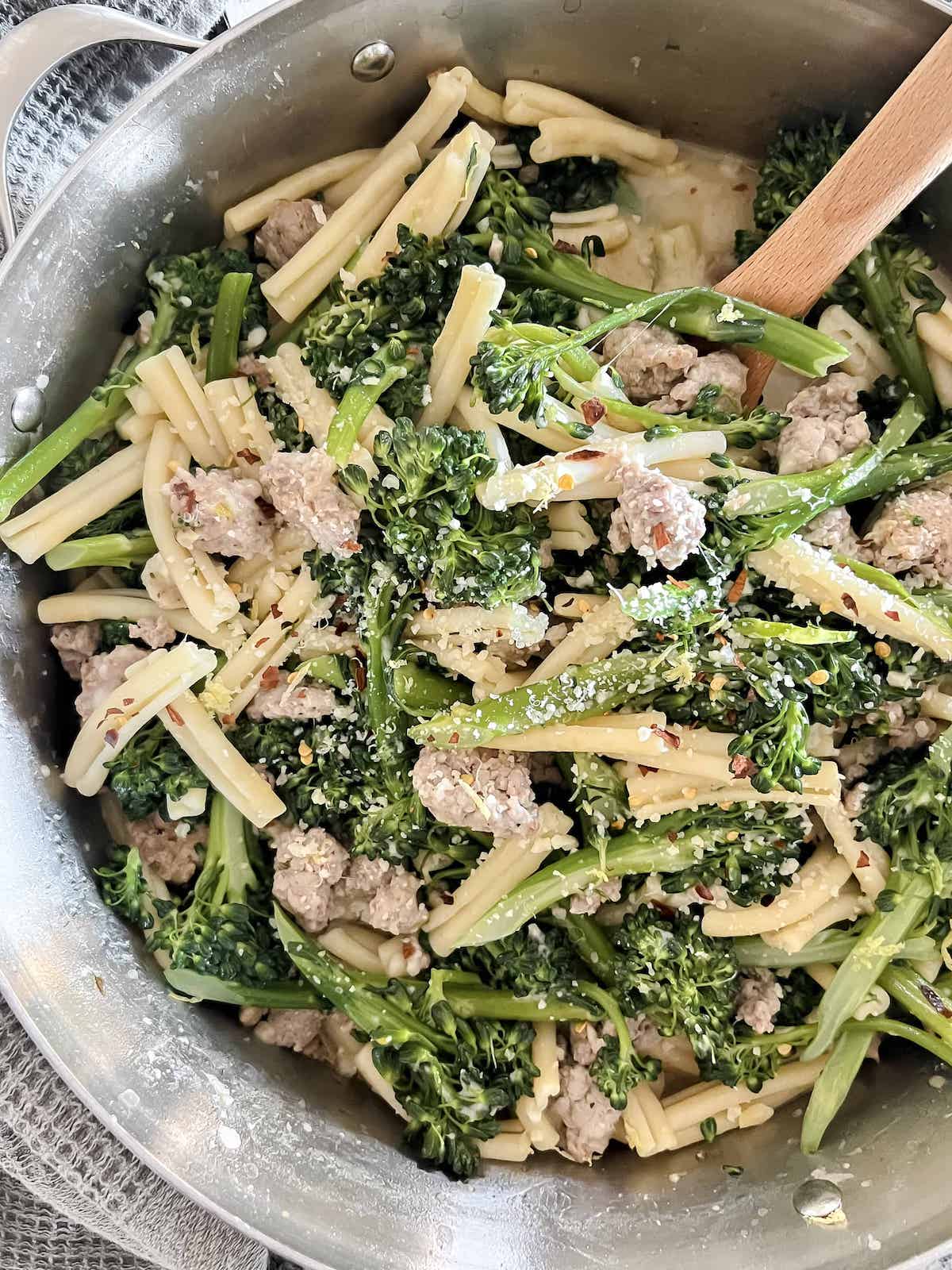 Silver pot full of casarecce pasta with broccolini and sausage with a wooden spoon.