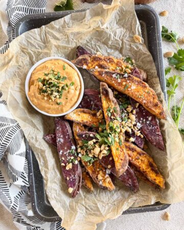 Quarter baking sheet with crumpled parchment paper with purple and orange sweet potato wedges topped with crushed peanuts and cilantro and a small white bowl of dipping sauce.