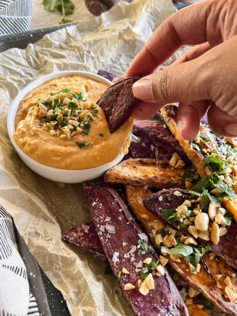 Crumpled parchment paper with sweet potato wedges topped with crushed peanuts and cilantro and a brown hand dipping a wedge into a small white bowl of peanut dipping sauce.