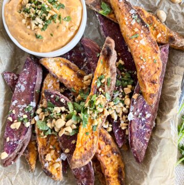Close up of mixed color sweet potato wedges on crumpled parchment paper topped with chopped peanuts and cilantro with a small white bowl of peanut dipping sauce.
