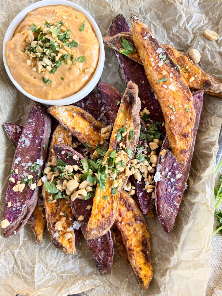 Close up of mixed color sweet potato wedges on crumpled parchment paper topped with chopped peanuts and cilantro with a small white bowl of peanut dipping sauce.