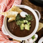 White bowl with small handles full of spicy black bean soup, topped with sour cream, diced avocado, cilantro and tortilla chips. Pink patterned napkin, cilantro and tortilla chips in the background.