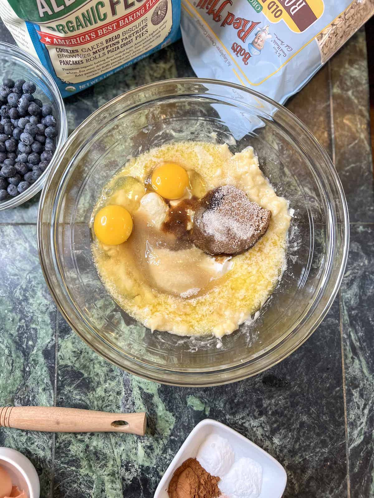 Adding wet ingredients to mashed bananas in a glass bowl.