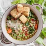 Grey bowl full of vegetable orzo soup topped with croutons and parmesan cheese. Fresh celery leaves and a towel in the background.