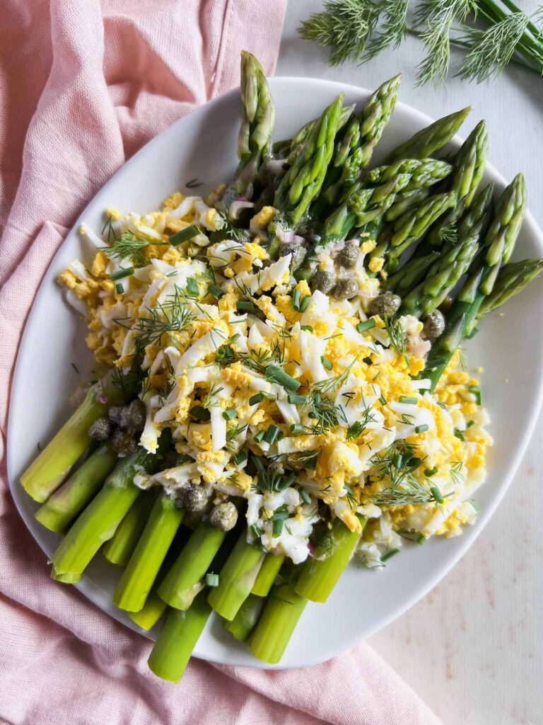 Asparagus spears on a white platter topped with grated hard boiled eggs and chopped fresh herbs with a pink napkin in the background.