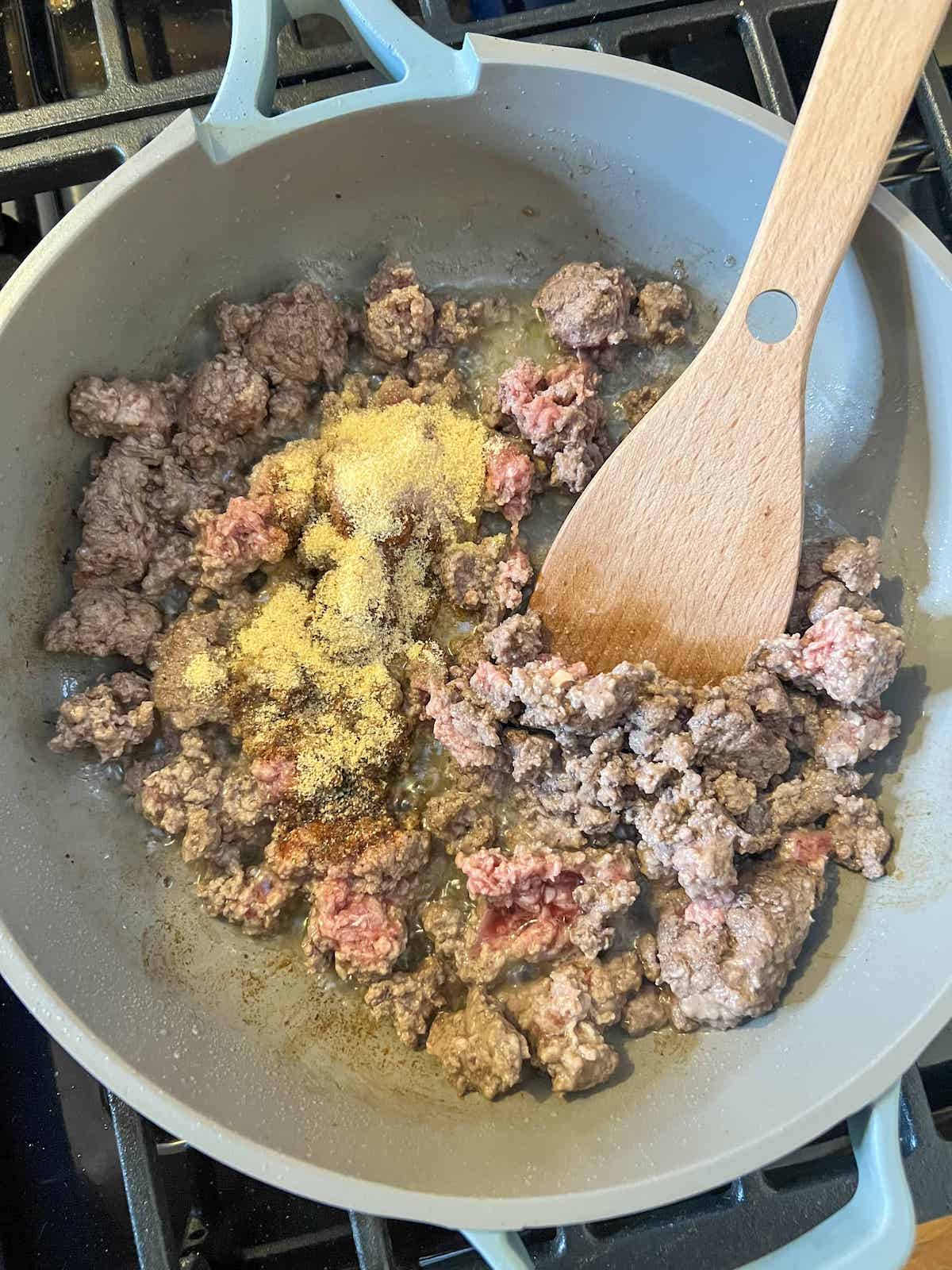 Ground beef cooking in a skillet with seasonings and a wood spatula.