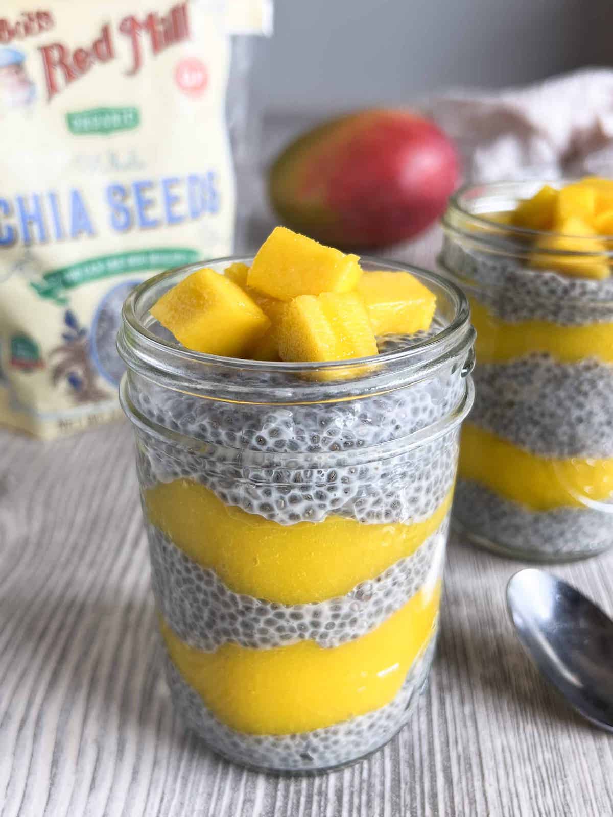 Chia pudding and mango puree layered in a mason jar with fresh mango chunks on top. Mango and bag of Bob's Red Mill chia seeds in the background.