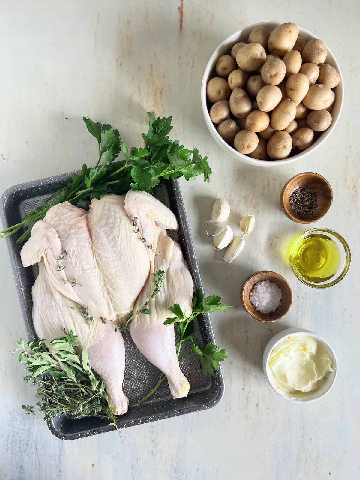 Ingredients for whole roast chicken and potatoes. Whole raw chicken on a tray with fresh herbs, white bowl of baby potatoes, garlic cloves, salt, pepper, oil and butter in little bowls.