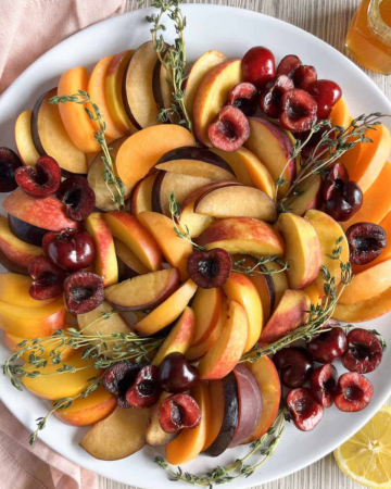 Sliced mixed stone fruit on a white platter with sprigs of fresh thyme.
