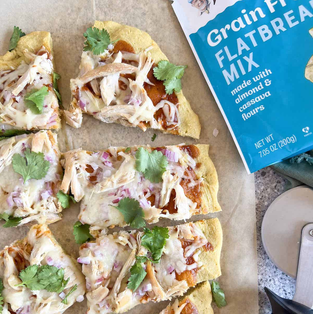 Slices of bbq chicken flatbread pizza on parchment next to a bag of Bob's Red Mill Grain Free Flatbread Mix and a pizza cutter.