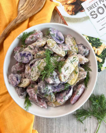 White bowl of purple and white potato salad with a wooden spoon on top of a yellow napkin, fresh dill sprigs and the Sweet Potato Soul cookbook cover in the background.