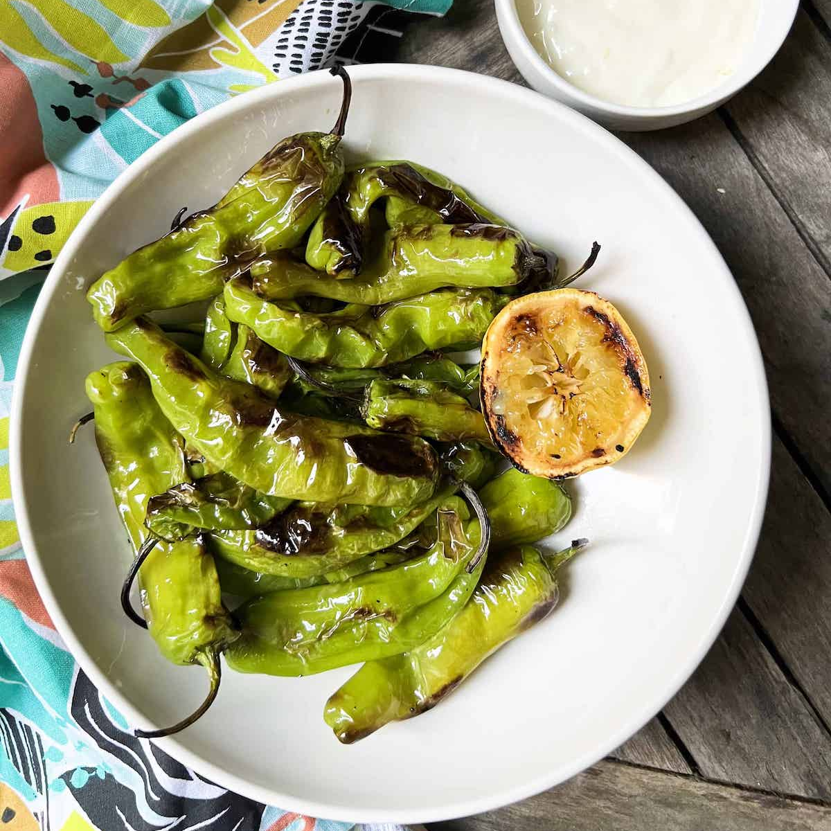 Bowl of charred shishito peppers with a small white bowl of aioli and a colorful printed napkin in the background.