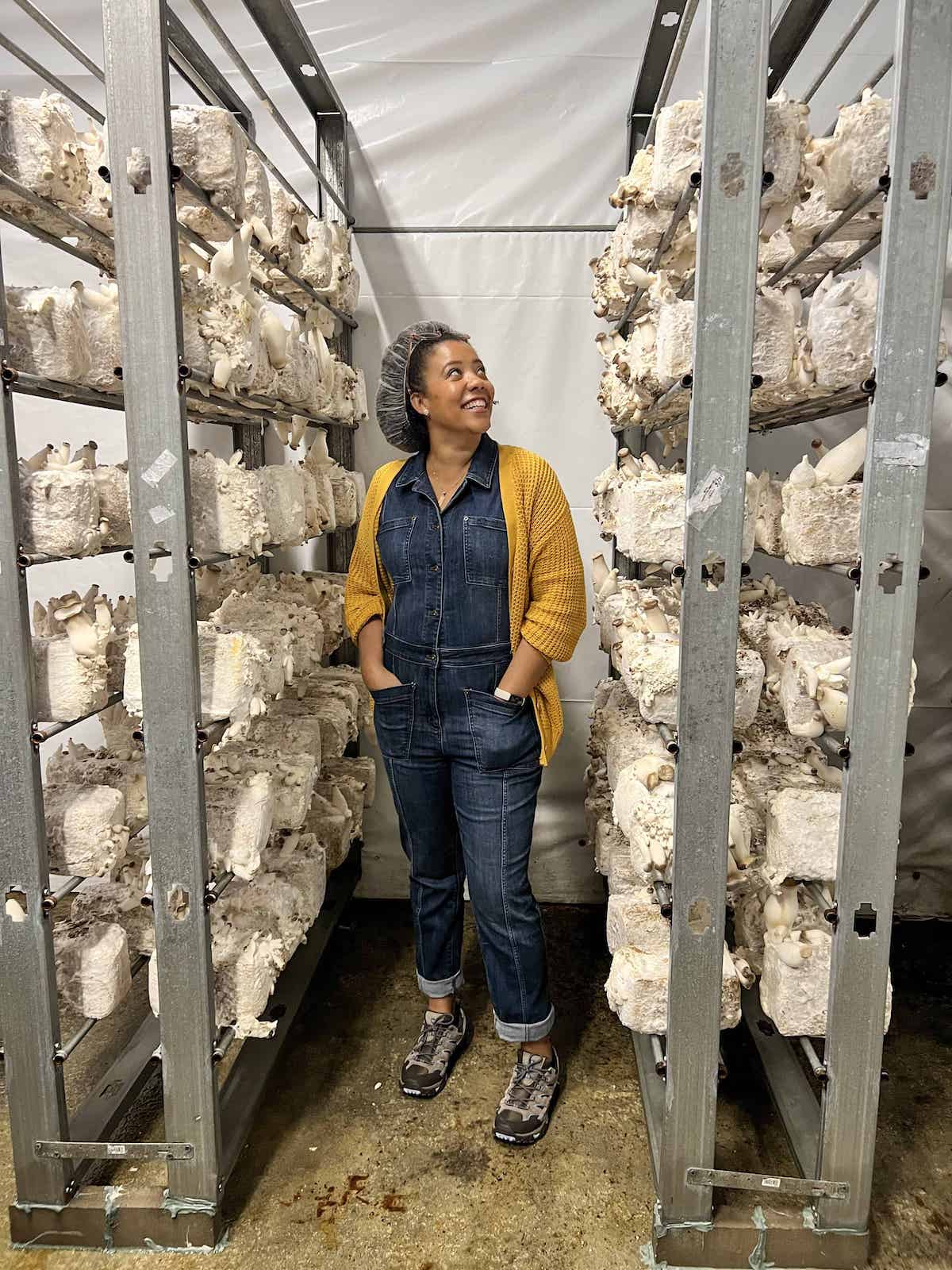 Jessica from Big Delicious Life standing between racks of mushrooms in the cultivation room at Far West Fungi.
