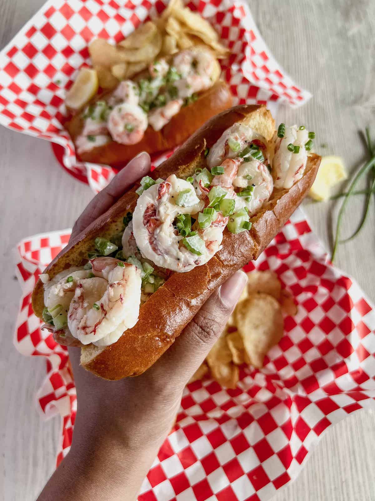 Brown hand holding up argentine red shrimp roll with deli baskets lined with red and white checkered paper below.
