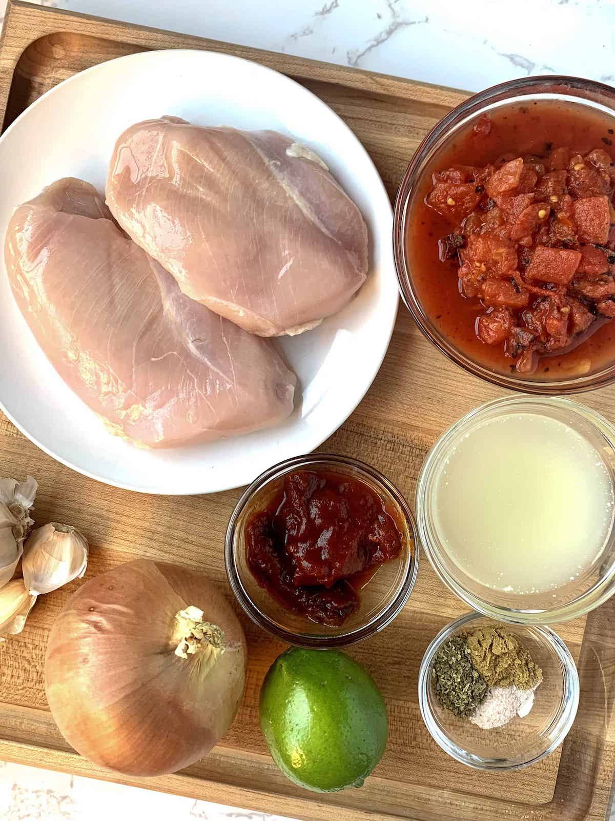 Ingredients for chicken tinga in small bowls on a wood cutting board with a white plate of raw boneless skinless chicken breasts.