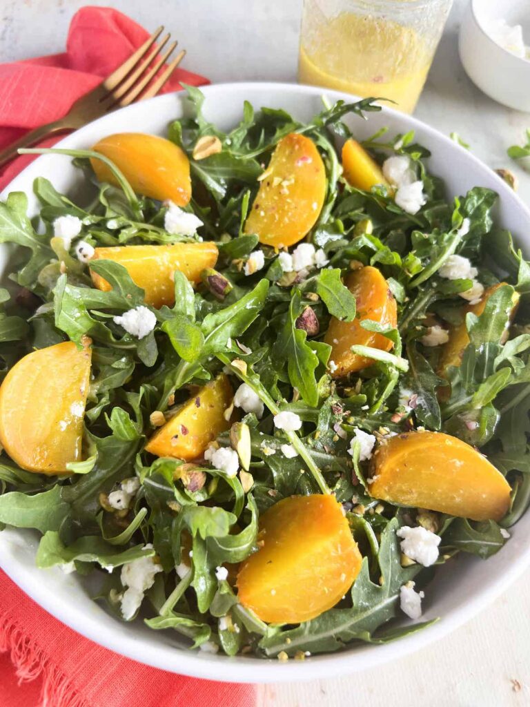 Golden beet and arugula salad with goat cheese and pistachios in a white bowl with a pink napkin under it. Mason jar of vinaigrette and bowl of goat cheese in the background.