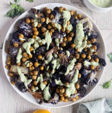 Roasted purple cauliflower and chickpeas drizzled with lemon herb tahini sauce in a white bowl.