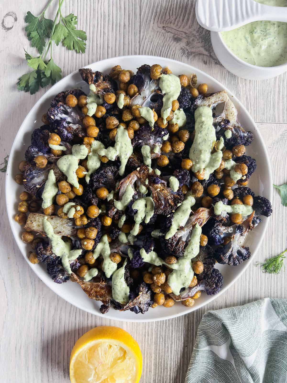 Roasted purple cauliflower and chickpeas drizzled with lemon herb tahini sauce in a white bowl.