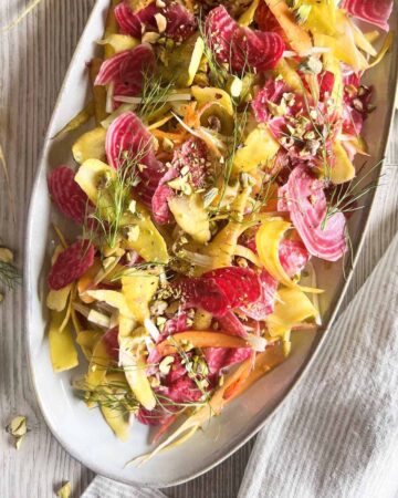 Shaved vegetable salad on a grey oval platter with yellow carrots and chopped pistachios on the table.