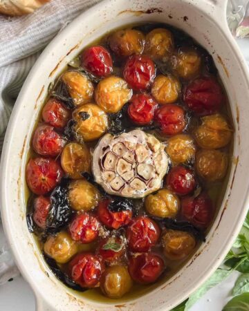 Oval baking dish with confit cherry tomatoes and a whole head of roasted garlic with herbs.