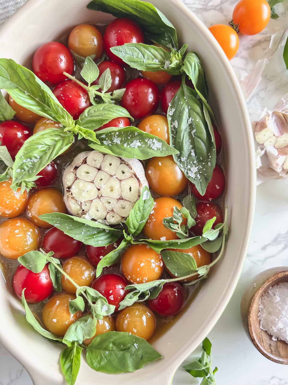Oval baking dish with raw cherry tomatoes, olive oil, a whole head of garlic with the top cut off and fresh herbs.