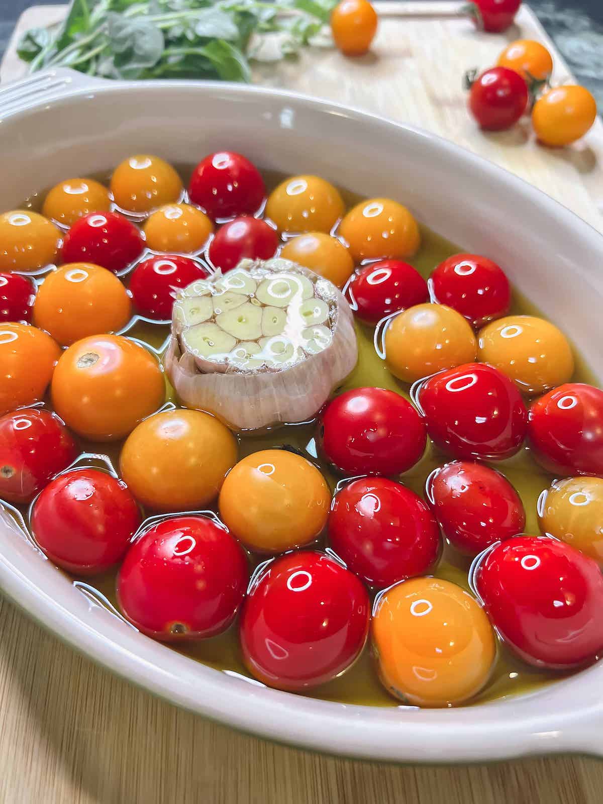 Oval baking dish full of cherry tomatoes with a whole head of cut garlic nestled in the middle and filled with olive oil.