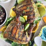 Two grilled mojo chicken leg quarters on a platter with fresh cilantro, lime and orange pieces with a bowl of rice, bowl of beans and bowl of marinade on the side.