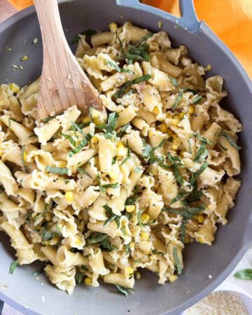 Skillet with creamy corn and basil pasta and a wooden spoon.