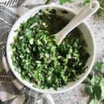 Small white bowl full of cilantro chimichurri sauce with a white spoon in it. Black and white patterned napkin and cilantro sprigs in the background.