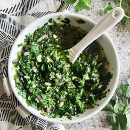 Small white bowl full of cilantro chimichurri sauce with a white spoon in it. Black and white patterned napkin and cilantro sprigs in the background.