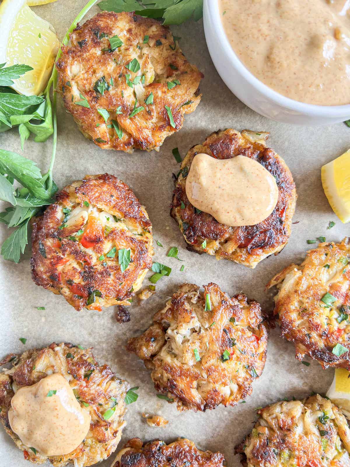 Crab cakes, garnished with parsley, on parchment paper with a dollop of remoulade sauce on one. 