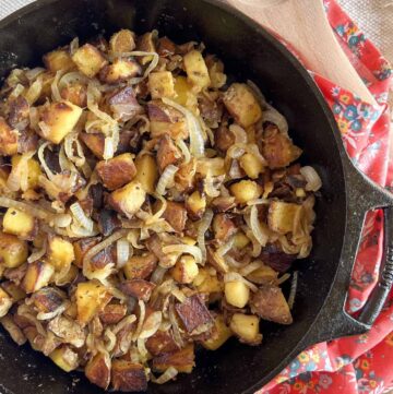 Fried potatoes and onions in a cast iron skillet with a wooden spoon and pink floral napkin in the background.