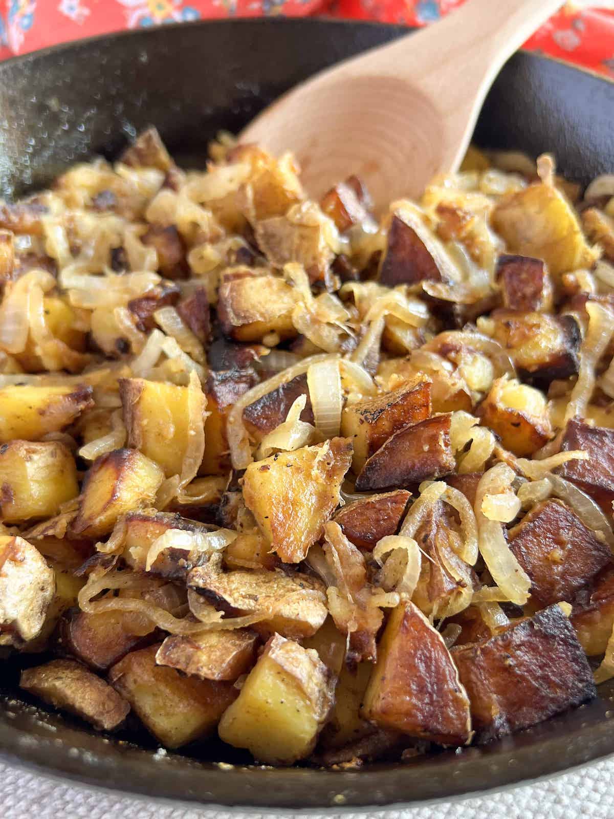 Close up of crispy pan fried potatoes and onions in a cast iron skillet with a wooden spoon.