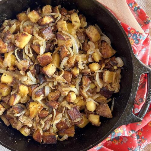 Fried potatoes and onions in a cast iron skillet with a pink floral napkin in the background.