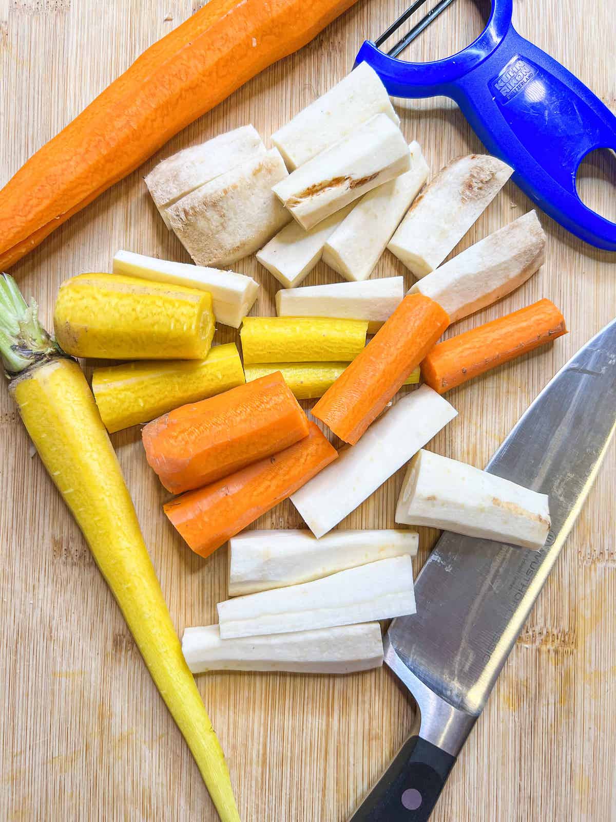 Cut carrots and parsnips on a wood cutting board with a knife and a blue vegetable peeler.
