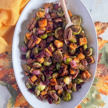 Roasted brussels sprouts and sweet potatoes topped with dried cranberries and pecans on a white oval platter with a wooden spoon on top of a leaf placemat and yellow napkin.