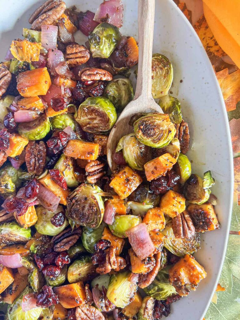 Roasted Brussels sprouts and sweet potatoes topped with dried cranberries and pecans on a white platter with a wooden spoon.