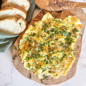 Wood board spread with softened butter, roasted garlic paste, herbs, flaky salt and drizzled with honey. Bread in a green napkin in the background.