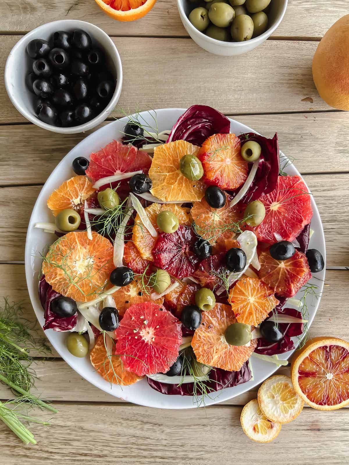 Round slices of mixed citrus fruit layered with thinly sliced fennel and topped with olives on a white plate on a wood table.