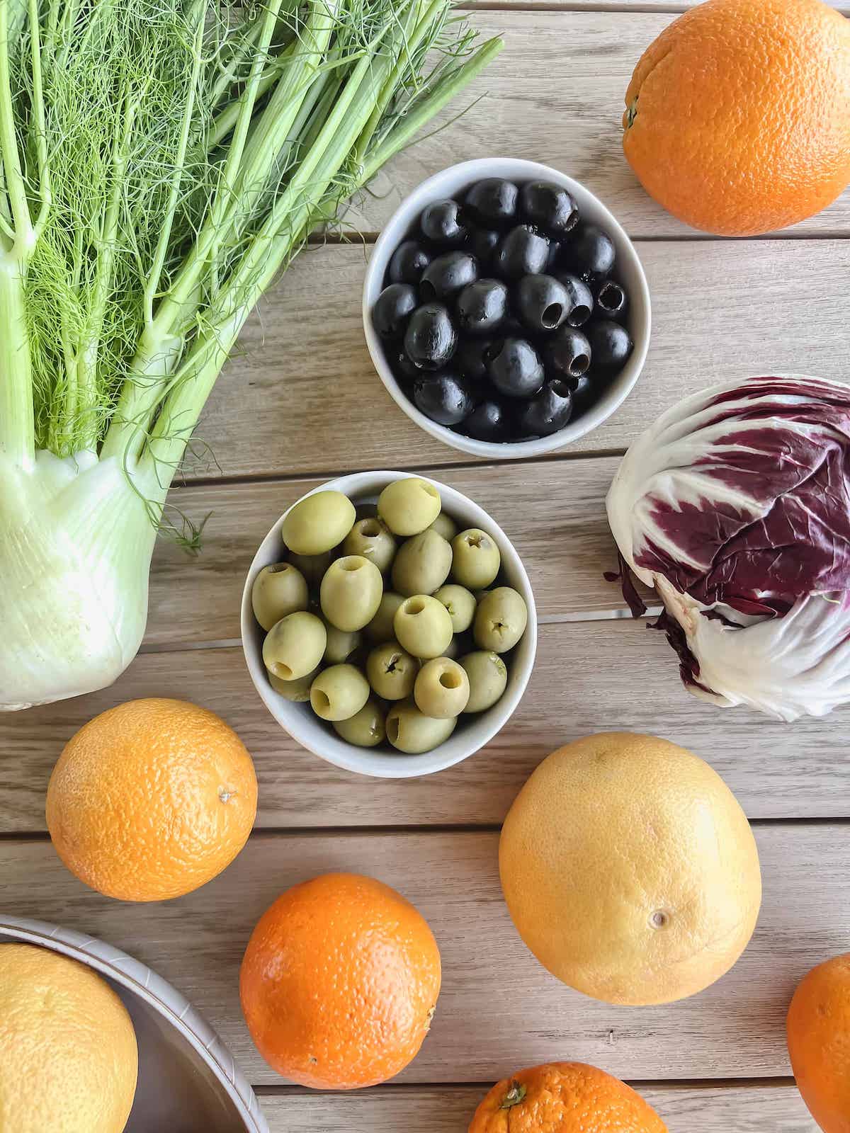 Two bowls of olives, mixed citrus fruits, fennel and a head of radicchio on a wood table.