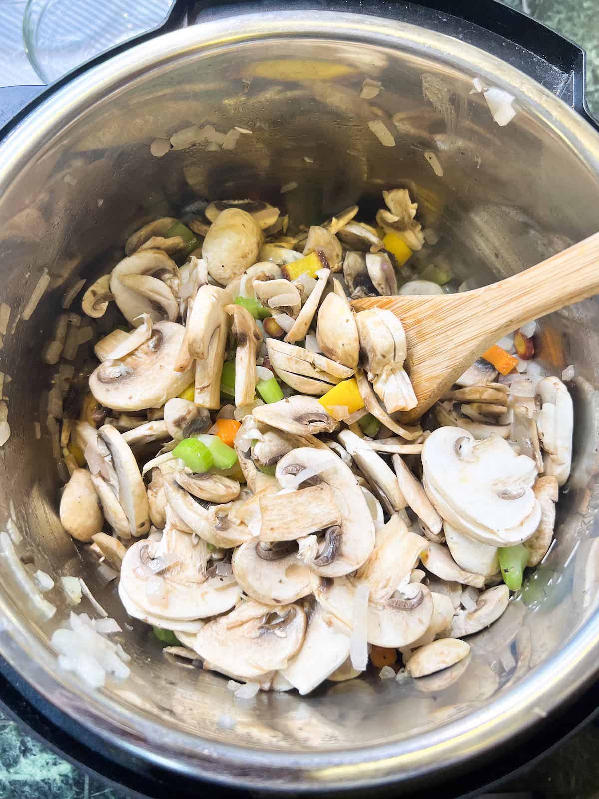 Sliced mushrooms and sauteed aromatics in an instant pot with a wooden spoon