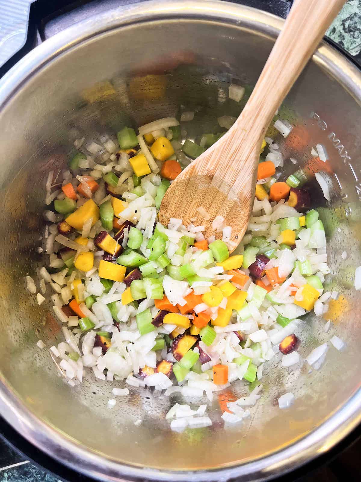 Diced onions, carrots and celery cooking in an Instant Pot with a wooden spoon.