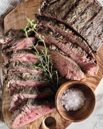 Sliced medium rare tri tip roast on a wood cutting board garnished with a sprig of thyme and a small wood bowl of flaky salt.