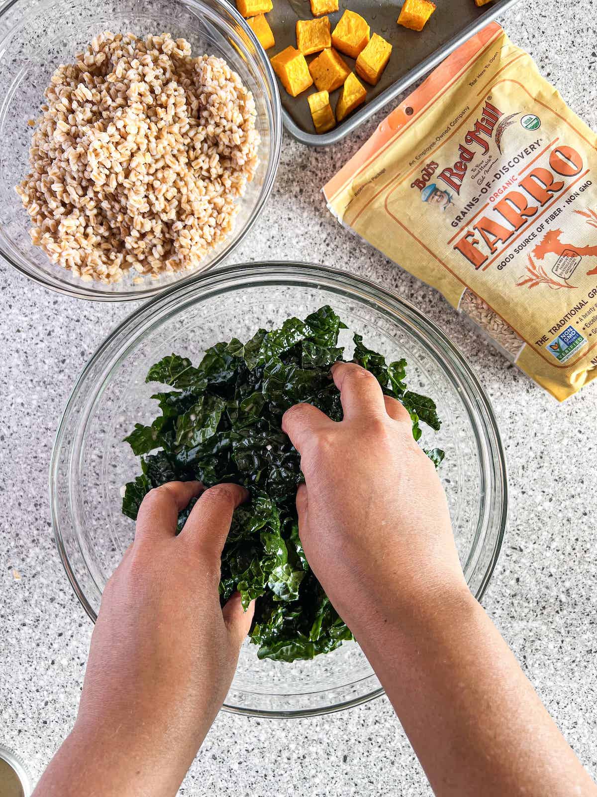 Brown hands massaging kale in a glass bowl.