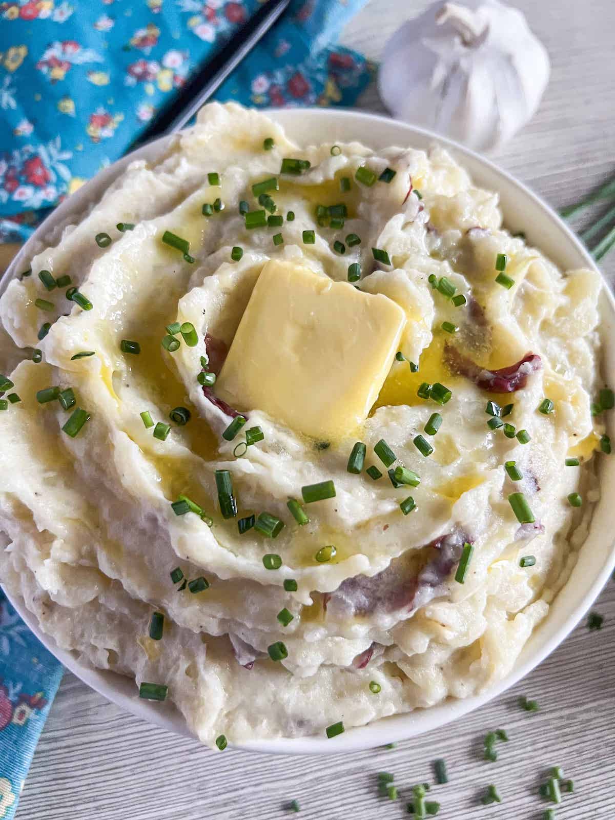 Bowl of red skin mashed potatoes topped with chopped chives and a pat of butter with a blue floral napkin in the background.