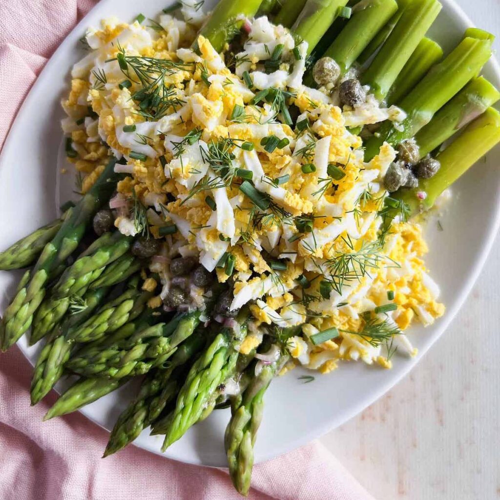 Asparagus spears topped with grated egg, fresh herbs and caper vinaigrette on a white plate.