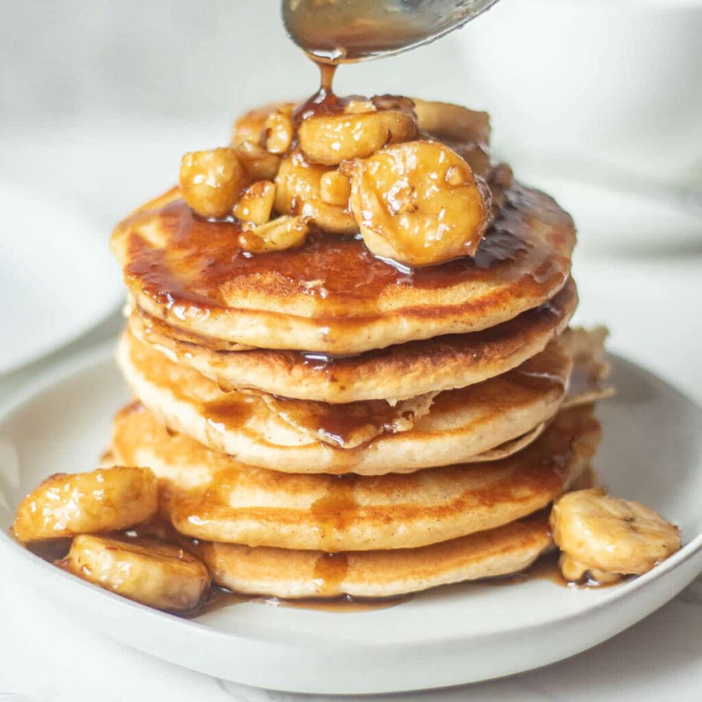 Stack of pancakes with bananas foster topping being drizzled with syrup.