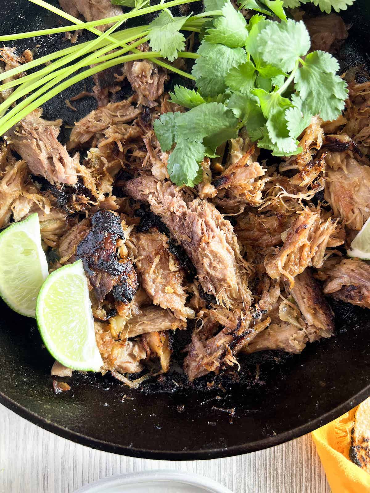 Close up of crispy pork carnitas in a cast iron skillet, garnished with cilantro and lime wedges.